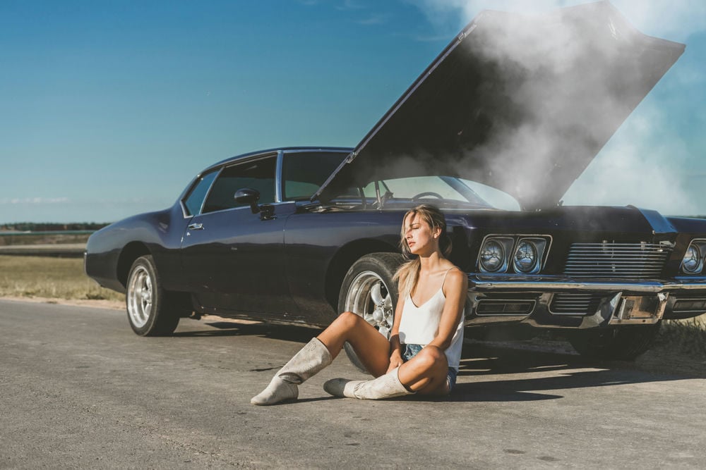 A woman waiting on the side of the road with an overheating engine.